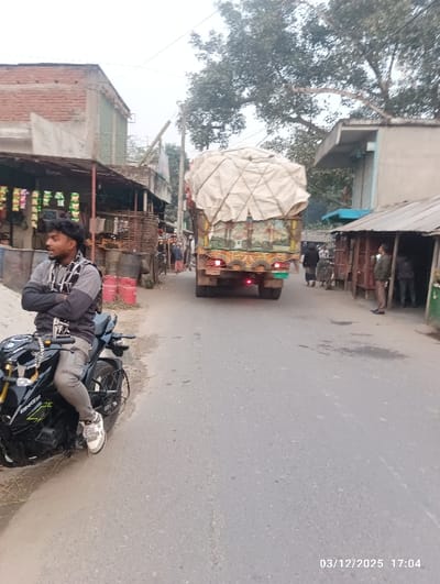 The image depicts a street scene in Maliat, Bangladesh, captured on March 12, 2025, at 17:04. In the foreground, a male individual with dark, curly hair and a mustache is seated sideways on a black motorcycle, identified as a Yamaha FZ-S, facing left. He wears a grey long-sleeved shirt with a patterned scarf around his neck and light-colored pants with white and grey athletic shoes. His arms are crossed.
A large, heavily laden truck, covered with a beige tarp, occupies the center of the asphalt road, moving away from the camera. The truck's rear exhibits colorful decorative painting and illuminated red taillights.
On the left side of the road, several multi-story buildings constructed from brick and concrete are visible. One ground-floor establishment displays packaged goods, likely snacks, hanging in front. Red gas cylinders are positioned near this building. Further down the street on the left, additional individuals are visible.
On the right side of the road, single-story structures, possibly made of corrugated metal or wood, line the street. Multiple individuals are standing outside these structures. The background features trees with sparse foliage and an overcast sky. Overhead utility lines are present across the scene.