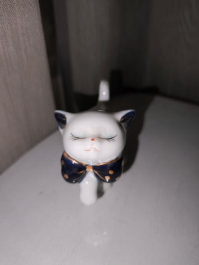A close-up, eye-level, static shot features a white ceramic figurine of a cat with its eyes closed, appearing to be in a state of peaceful repose. The cat figurine is adorned with a dark blue polka-dot bowtie with gold accents around its neck. Its ears are also tipped with dark blue. The figurine is positioned on a smooth, light-colored surface that may be a tabletop or shelf. The background consists of dark, indistinct forms, possibly curtains or shadows, creating a muted and slightly shadowed environment. The lighting in the scene is dim, with some light reflecting off the glossy surface of the figurine, particularly on its face and bowtie. There are no people or other animals visible in the image. The overall impression is of a quiet, indoor setting, focusing solely on the decorative ceramic cat.