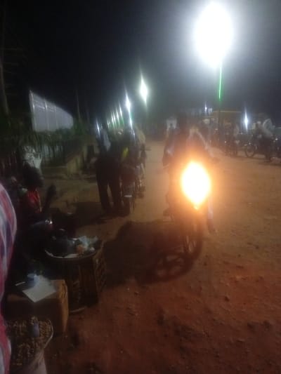 This is a nighttime photograph taken in Tundun Wada West, Nigeria, depicting a street scene. The image is illuminated by a bright, overhead street lamp and the headlight of a motorcycle in the foreground. Several people are visible, some standing and others on motorcycles. In the lower left of the frame, a person is seated behind a display of what appears to be food items in baskets. The ground is unpaved, consistent with a rural or semi-urban area. A white, translucent structure, possibly a fence or wall, is visible on the left side of the image, with vegetation or trees in front of it. The overall atmosphere is that of a bustling night market or gathering place.