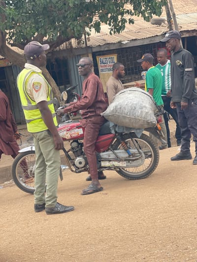 In Chikaji, Nigeria, a group of men stand near a motorcycle heavily laden with a large sack-like object. One man, in a yellow safety vest, holds a long black object near the motorcycle. The other men are dressed in casual attire. In the background, there's a building with a sign that includes the words "SALES GIRL Needed Urgently". The street appears to be paved.