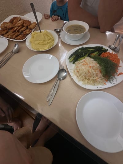 A table is laden with food and dinnerware, set for a meal. In the foreground, a white oval plate is empty. To the left, a white oblong plate is filled with golden mashed potatoes. Next to it, a white square platter holds an assortment of fried cutlets. In the center of the table, a white bowl of soup with visible mushrooms sits on a saucer. To the right, a large white oval plate is piled with sauerkraut, shredded carrots, sliced cucumbers, and dill. Several spoons and forks are scattered across the table, alongside additional white plates. In the background, a child wearing a blue shirt is visible, with their hands resting on the table. Parts of an adult's arms and legs are also visible, suggesting multiple people are seated at the table. The setting appears to be indoors, likely a dining area. The image context suggests this is likely a domestic scene within the Завалочыцкі сельскі Савет region of Belarus.