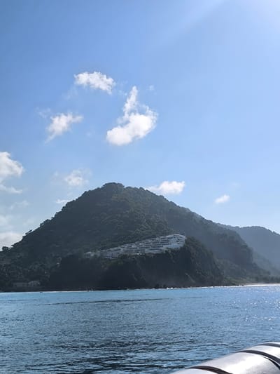 A bright, sunny day illuminates a coastal scene in Karangasem, Indonesia. The image captures a wide expanse of deep blue ocean, with small waves creating a shimmering surface due to sunlight. In the distance, a lush, tree-covered mountain slopes down towards the sea. Perched on the hillside is a modern resort or housing complex with white buildings featuring multiple levels and balconies. The sky above is a clear, bright blue, dotted with a few wispy white clouds. The sun's rays are visible, casting a bright glow and creating a slightly hazy effect in the distance. The perspective suggests the photo was taken from a boat or elevated viewpoint on the water, looking towards the shore.
