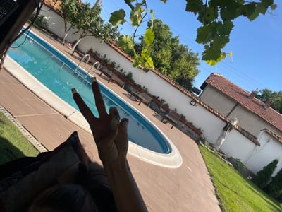 A first-person perspective shot shows a hand making a peace sign in front of a swimming pool in a backyard. The hand has long, light-colored nails with a metallic sheen. The swimming pool is oval-shaped with bright blue water and a white edge. The pool area is paved with light brown tiles, and there is a strip of green grass to the left of the pool. In the background, a white wall runs along the property line, with red-tiled roofs of houses visible above it. There are trees and some lounge chairs on the other side of the pool. The sky is clear and blue. The image appears to be taken in Totleben, Bulgaria, based on the reference to the city.