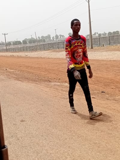 The image displays a male subject walking on a paved surface in Katcha, Nigeria. The subject, appearing to be a young adult, possesses a dark complexion and short, dark hair. He is attired in a long-sleeved shirt featuring a multi-colored tie-dye pattern primarily in red, yellow, black, and purple, with a graphic design on the chest. He wears black trousers and light-colored sandals. The subject is oriented with his body angled slightly towards the right side of the frame, moving generally leftward. To the subject's right is an unpaved area of reddish-brown earth. The background features a low concrete block wall or fence spanning horizontally, behind which utility poles with overhead lines and sparse trees are visible. The sky is hazy or overcast.