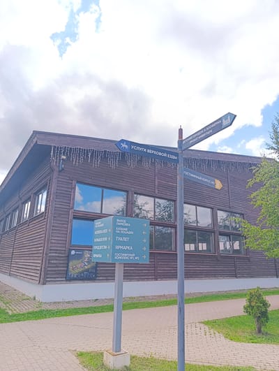 The image shows a wooden building with a signpost in front of it, located in the city of Chernogolovka, Russia. The signpost has multiple directional signs pointing to various locations. One sign indicates "HORSEKA KIDS", "BIG FIELD", "CHILDREN'S WORLD", and "FAIR". Another sign points to "RIDING ON HORSES", "TOILET", and "FAIR". A third sign states "HOTEL COMPLEX №1, №2". A blue sign with a horse icon points towards "HORSE RIDING SERVICES". Further up, a sign in Russian reads "CHILDREN'S TOWN AND ZOOFERMA" and "INVENTORY RENTAL". The building appears to be a recreational facility, possibly a horse riding center or a park. The environment is outdoors, with paved walkways, grass, and trees. The sky is partly cloudy.