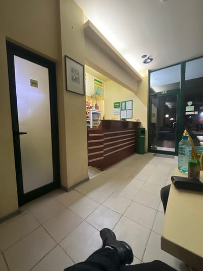 The image shows the interior of a building in Nova Zagora, Bulgaria. The space appears to be a commercial establishment, possibly a restaurant or cafe. There is a reception desk, a WC door, and the entrance is visible, leading to the outside. A framed picture hangs on the wall. The flooring is tiled. The presence of the WC indicates customer use. There is a person partially visible in the image.