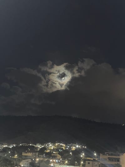 This is a nighttime photograph taken from a slightly elevated vantage point, looking over a city that appears to be Tiberias, Israel. The foreground is dominated by the dark silhouette of a hilly landscape, likely Mount Arbel, with a town spread out at its base. The town is illuminated by numerous streetlights and building lights, creating a scattered, golden glow against the dark backdrop.
The sky above is a deep, dark blue, with a prominent moon partially obscured by dramatic, dark clouds. The clouds have an interesting formation, with the moon appearing to be nestled within a heart-shaped opening. The moonlight illuminates the edges of the clouds, giving them a soft, ethereal glow. There are a few faint stars visible in the sky, scattered among the clouds. The overall impression is one of a serene, romantic night scene over a populated area.