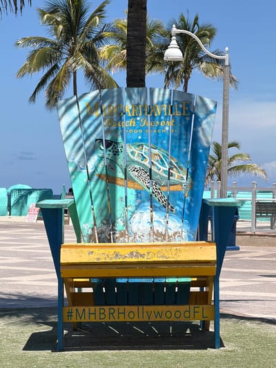 The image shows a brightly colored adirondack chair with a painting of a sea turtle on its backrest. The chair is located outdoors, likely at a beach or resort, given the palm trees and the bright blue sky. The chair itself is painted in vibrant blues and greens, with the sea turtle depicted in a detailed, lifelike manner. The chair has yellow wooden armrests.

Visible text on the chair includes "MARGARITAVILLE Beach Resort Hollywood, FL" on the backrest, and "#MHBRHollywoodFL" vertically on the right side of the chair's frame. The setting appears to be a sunny day, with clear blue skies and dappled sunlight casting shadows from palm fronds onto the chair and the ground. The ground in front of the chair is a light-colored paved area, and to the right, there is a patch of green turf. In the background, there are other beach chairs and tables, suggesting a public area designed for relaxation and leisure. The overall impression is one of a tropical, vacation-like atmosphere.