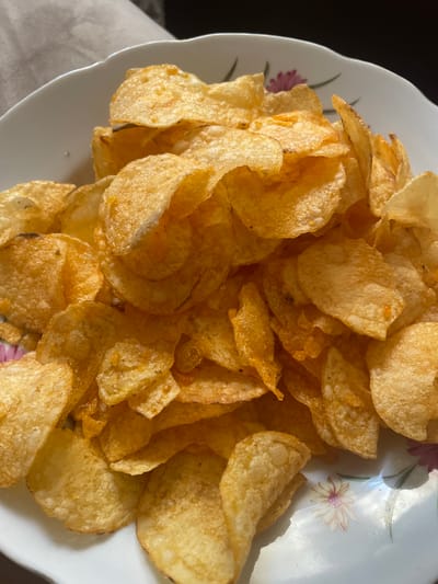 A close-up, high-angle shot shows a white ceramic bowl filled with golden-brown potato chips. The bowl has a decorative rim with a subtle floral pattern featuring pink and purple flowers with green stems. The chips are varied in shape and size, with many showing the characteristic crinkled texture of kettle-cooked chips. Some chips are whole, while others are broken into smaller pieces. The lighting casts shadows across the chips, highlighting their texture and creating a sense of depth. The background is dark, drawing attention to the food. The overall impression is one of a simple, appetizing snack. There is no visible text in the image.