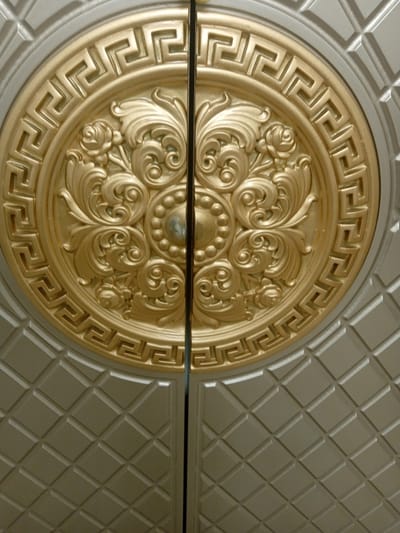 The image shows a close-up, low-angle view of ornate, decorative doors. The doors appear to be made of metal and feature a prominent circular gold medallion with intricate relief patterns. This medallion has a Greek key border surrounding a central floral design with rose motifs and swirling scrollwork. The remainder of the doors is a matte silver color with a diamond-shaped embossed pattern. The doors are split down the middle by a vertical dark line, suggesting they are a pair. There is no indication of the location in Unkurgan, Uzbekistan, or any people present. The scene is static and focused on the detailed craftsmanship of the doors.