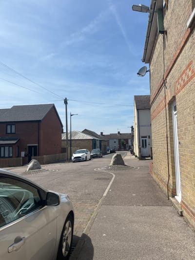 This image depicts a residential street in East Suffolk, United Kingdom. The view is taken from street level, looking down the road. The street is narrow, flanked by brick buildings on both sides. One side of the street has a sign reading "Cambridge Street". A silver car is partially visible on the right side of the frame, parked along the street. Further down the street, other parked cars are visible. The road surface is asphalt, marked with white lines. Periodically, there are raised stone planters at the road side with what appears to be wireframe surrounding. Overhead, power lines run along the street, supported by utility poles. The sky is blue with scattered clouds. A surveillance camera and a satellite dish are mounted on the side of one of the buildings.