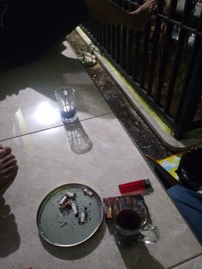 The image depicts an outdoor scene at night, likely on a tiled patio or balcony. In the foreground, a collection of used cigarette butts rests in a round, metallic ashtray. Adjacent to the ashtray are a pack of cigarettes, a red lighter, and a glass mug filled with a dark liquid, possibly coffee. To the left, a person's bare feet are visible on the tiled ground. In the background, a metal railing and a narrow drainage channel can be seen. A pair of sandals is placed near the drainage channel. The overall lighting suggests it is nighttime, with artificial light casting shadows on the tiled surface. The context suggests a moment of relaxation or a break, likely in a residential area in Sukamanah, Indonesia.