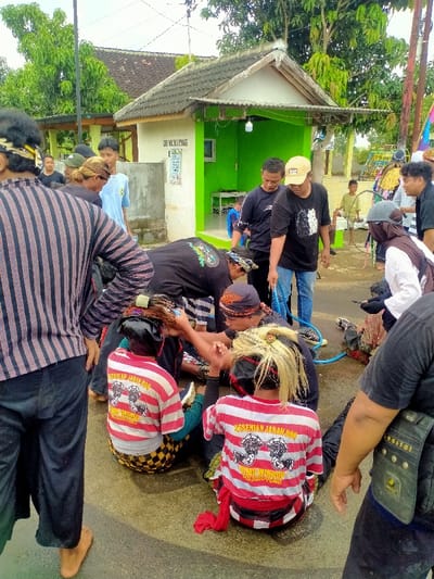 The scene takes place in a residential area in Kecamatan Kedawung, Indonesia, at the coordinates -7.52 latitude and 111.029 longitude. A group of people are gathered around a man in a white and red striped shirt with the words "Kesehian Jarak, 0000000000" on it. The man is lying on the ground in front of a small building with green painted walls. Two other men in black shirts appear to be aiding him. There are multiple people around them, including one man in black and white striped shirt, several other men in black shirts, and a man in a white shirt with a red and black plaid scarf. There is a blue hose on the ground.  The scene appears to be taking place in a street, with a small building and other residential structures visible in the background.