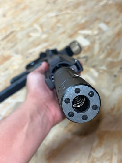 A close-up, slightly angled shot shows a person's hand holding a firearm accessory, possibly a suppressor, with a wooden background. The accessory is dark gray with six small black spherical objects arranged in a circle around a central opening. Inside the opening, a coiled spring is visible. The hand, which appears to be a light skin tone, is holding the accessory from behind, with fingers wrapped around the main body of the firearm. The background is composed of light brown, textured material, likely unfinished wood, with visible grain patterns. The focus is sharp on the firearm accessory, while the hand and background are progressively blurred. No specific location information for Vasilyevo, Russia is discernible from the image.