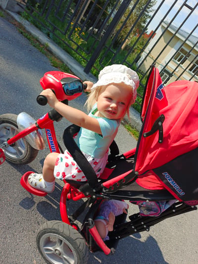 A young Caucasian girl wearing a white bonnet and a light blue t-shirt with a cherry-patterned skirt is seated on a red tricycle with a stroller attachment. She is holding the handlebars of the tricycle, which has a silver fender and a red frame with "FORMUL" visible on it. The stroller is bright red and black with "FORMULA 3" printed on it. A doll is visible inside the stroller. The scene takes place outdoors on an asphalt surface, with a black metal fence and a building in the background. The image appears to have been taken in the city of Zlatoust, Russia, indicated by the presence of a unique black metal fence often seen in this region. The lighting suggests it is daytime with bright sunlight.