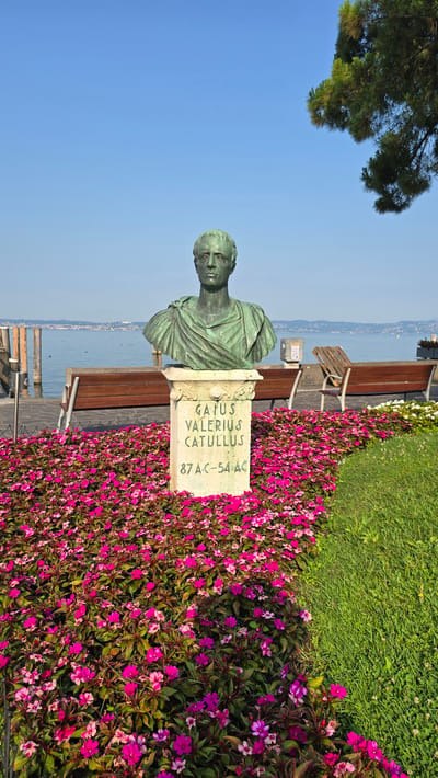 A bronze bust of Gaius Valerius Catullus, an ancient Roman poet, stands on a stone pedestal surrounded by vibrant pink and purple impatiens flowers. The bust is positioned in the foreground, with the inscription "Gaius Valerius Catullus 87 a.C.-54 a.C." clearly visible on the pedestal. Behind the floral display, a paved walkway runs alongside a body of water, likely Lake Garda, with wooden benches and pilings visible. In the background, a distant shoreline with buildings and hills can be seen under a clear blue sky. A pine tree branch overhangs the scene from the upper right. The overall setting suggests a park or promenade in Sirmione, Italy, dedicated to the poet.