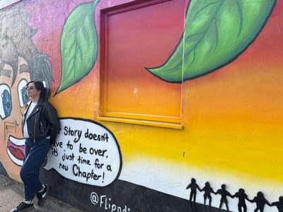 A woman is standing in front of a colorful mural located in Gorleston-on-Sea, United Kingdom. The mural features a cartoon face on the left, two large green leaves, a window painted with red and orange hues, a speech bubble that reads "Story doesn't have to be over, it's just time for a new Chapter!" and at the bottom are silhouettes of people holding hands. The mural's background consists of gradient colors ranging from pink and orange at the top to yellow and white at the bottom. The woman is wearing sunglasses, a black leather jacket, a white t-shirt, and blue jeans, and is leaning against the mural.