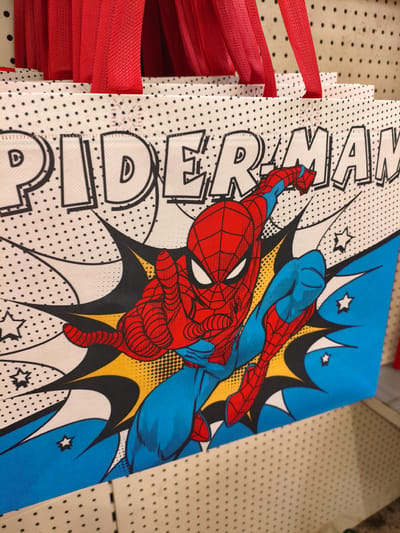 The image shows a close-up of several red fabric shopping bags with the Spiderman comic book design. The bags have white handles and are stacked vertically. The design features a large, dynamic image of Spiderman in action against a background of comic book-style sound effects and a dotted pattern. The image is cropped to focus on the bags and their design, with some of the red handles visible at the top. The background is a pegboard, typical of a retail environment. The location is not specified in the image or its metadata.