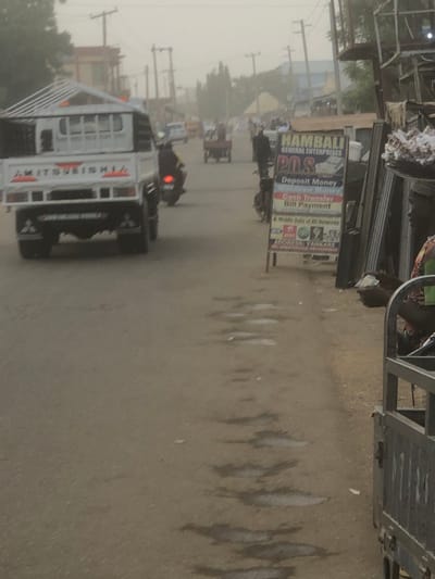 A hazy street scene features a Mitsubishi truck in the foreground. A sign for "HAMBALI GENERAL ENTERPRISES" advertises "POS, Deposit Money, Withdraw Money, Cash Transfer, Bill Payment" and "Mobile Data of All Networks." The address is listed as "YANKARE." A motorcycle and other vehicles are visible further down the road. Overlapping with other metal structures are what looks like merchandise or other metal parts.