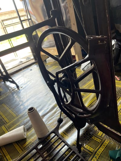 The image shows the underside of a vintage sewing machine. The machine's metal framework is visible, including a large, spoked wheel. Two spools of white thread are also present, one resting on the machine and the other on the patterned fabric beneath it. The fabric has a distinctive yellow and black geometric pattern. The background suggests an indoor setting, possibly a workshop or storage area, with some natural light filtering in from windows. The overall impression is of an old, well-used sewing machine, possibly in a rural Nigerian context, given the "Jauro Bamvo, Nigeria" location context. No people are visible in the image. The lighting suggests it could be daytime.