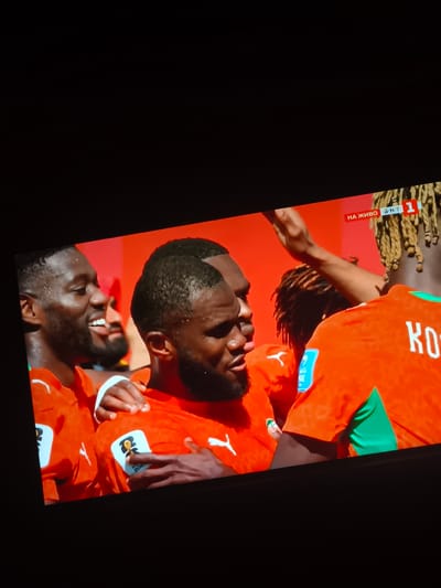 A group of Black male soccer players in orange jerseys are celebrating on a field. The player in the foreground on the left has a beard and is smiling widely. Behind him, another player with a beard is visible. To the right, a player with blonde braided hair and wearing an orange jersey is visible from the back, his arm raised. On his jersey, the word "KO" is visible in white letters. The jerseys also have a FIFA World Cup 2022 patch. In the upper right corner of the screen, a broadcast overlay shows "НА ЖИВО" (meaning "LIVE" in Bulgarian) and the number "1" within a red rectangle, indicating the broadcast originates from Harmanli, Bulgaria. The background is a solid red color.