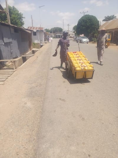 In Jimeta, Nigeria, on a sunny day, a person wearing a dark purple tunic and head covering pushes a trolley laden with yellow jerrycans filled with liquid. The trolley has a metal frame and two wheels. The jerrycans are arranged in two rows on the trolley, with their red caps clearly visible. To the right of the trolley, another person, possibly male, walks ahead, wearing a light-colored shirt and pants and a straw hat. The background shows a street scene with buildings on the left and right sides. On the left, there are corrugated metal structures and a drainage system. On the right, there are shops with signs, including one that appears to say "BARBER SHOP" and another that might be a "POS" station. Further down the street, a silver car and a yellow auto-rickshaw are visible. The overall scene suggests a typical day in a busy street market or commercial area, with people engaged in the transport of goods. The weather appears to be warm and bright, indicative of a daytime scene.