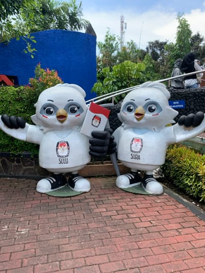 The image shows two white, cartoon-like figures of birds standing on a brick pathway. They are wearing red and white shirts that say "Komisi Pemilihan Umum" in black and "Sulu" and "Sura" in black underneath, respectively. The figure on the left is holding a red and white pamphlet with a logo in the center. The figure on the right is holding a stick. Both figures are facing forward with their arms extended outward. The background shows a blue wall with a partially visible brick wall behind it, green bushes, and a tree. The sky is partly cloudy. There is a sign in the background reading "Rute Selanjutnya" in black. The location of the photo, according to the coordinates provided, is a park in the city of Batu, Indonesia.