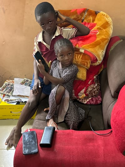 Two children, a boy and a girl, are seated on a couch in Gidan Waziri, Nigeria. The boy, on the left, is wearing a maroon and white collared shirt and blue shorts. He is holding a black smartphone. The girl, on the right, is wearing a patterned dress and is looking at the camera. They are both partially covered by colorful blankets. In front of the couch, on a red surface, are two black electronic devices, likely a smartphone and a power bank. To the left of the couch is a yellow box labeled "SOLAR FAN" and some papers. The wall behind them is a light orange color.