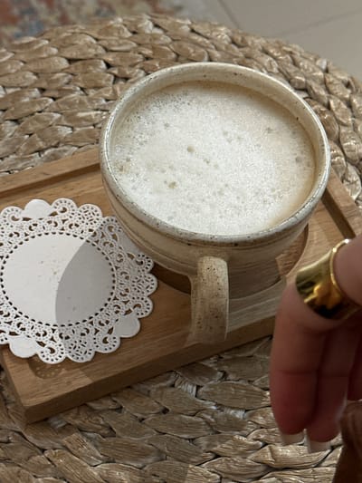 A high-angle shot shows a mug filled with a light-colored, frothy beverage, likely coffee or latte, sitting on a small wooden tray with a white doily underneath. A hand wearing a wide gold ring is partially visible in the upper right corner of the frame. The setting appears to be a cafe or home in Kerobokan Kelod, Indonesia, based on the background texture that resembles woven straw or rattan, common in the area. The objects are stationary, and there is no apparent interaction between the hand and the mug. The environment seems to be well-lit, suggesting daytime.