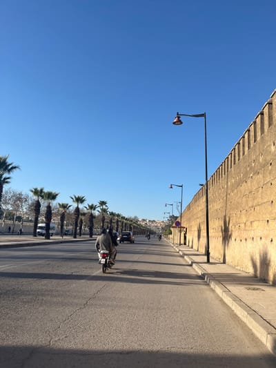 The image depicts a daytime street scene in Mechouar Fes Jdid, Morocco, characterized by a clear blue sky, a historic fortified wall, and a tree-lined boulevard.
On the right, a tall, light-brown, textured historical wall with battlements and small circular openings runs parallel to the road, extending into the distance. Shadows from unseen objects and streetlights are cast diagonally across its surface. A concrete sidewalk with a curb borders the wall. Several black streetlights with reddish, bell-shaped lamps are evenly spaced along the sidewalk. A red circular traffic sign with a white horizontal bar is visible on the sidewalk further down.
The center of the image is dominated by a multi-lane asphalt road, dry and showing minor surface cracks, receding into the background. A light-colored motorcycle with two individuals, the driver wearing a grey jacket and a dark helmet, and a passenger, is prominent in the foreground of the rightmost lane, traveling away from the viewer. Further down the road, additional motorcycles and at least one dark passenger car are also moving in the same direction.
On the left side of the road, a long, orderly row of slender palm trees with green fronds defines a median or an opposing traffic lane. Beyond the palm trees, a white van and another dark vehicle are visible, traveling in the opposite direction. In the far distance, indistinct buildings and structures rise on a hillside under the clear sky, forming a distant urban landscape. The scene is illuminated by natural sunlight, casting long shadows indicative of morning or late afternoon.