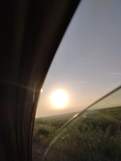 The image presents a striking view of a brilliant, overexposed sun, likely observed through the sunroof or angled window of a moving vehicle. The sun, a vivid circular light source, dominates the upper right-center of the frame, bathing the surrounding sky in a warm, hazy glow. The sky itself appears diffused, with faint, elongated streaks visible, which could be thin clouds or smudges/reflections on the glass.

The immediate foreground and lower left quadrant are occupied by the dark, textured interior material of the vehicle, possibly a car's headliner or window frame, providing a strong dark contrast to the bright exterior. To the far right, beyond what appears to be another window or opening, is a glimpse of dense, dark green foliage. This vegetation, likely trees or bushes, is somewhat blurred and indistinct, suggesting the vehicle is in motion.

The intense backlighting from the sun strongly indicates either sunrise or sunset, creating a dramatic atmosphere. The scene evokes a sense of travel or a journey through a natural landscape in Roza, Bulgaria. No people are visible, and no discernible text appears in the image.