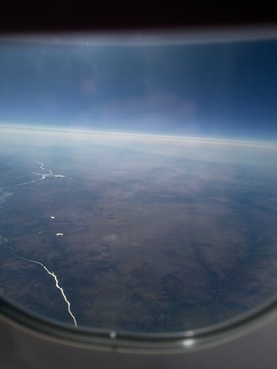The image is an aerial view of a vast, arid landscape seen through an airplane window. A winding, bright river or waterway cuts through the dark, textured terrain, reflecting the sunlight. Scattered across the land are smaller bodies of water or reflective patches. The horizon is blurred by atmospheric haze, with layers of clouds visible above the land, transitioning into a deep blue sky. The foreground is dominated by the dark, curved frame of the airplane window. There are no people or specific structures visible in the scene. The location is not identifiable as Santa Marta de Magasca, Spain, as the landscape appears rugged and sparsely populated, not typical of that region.