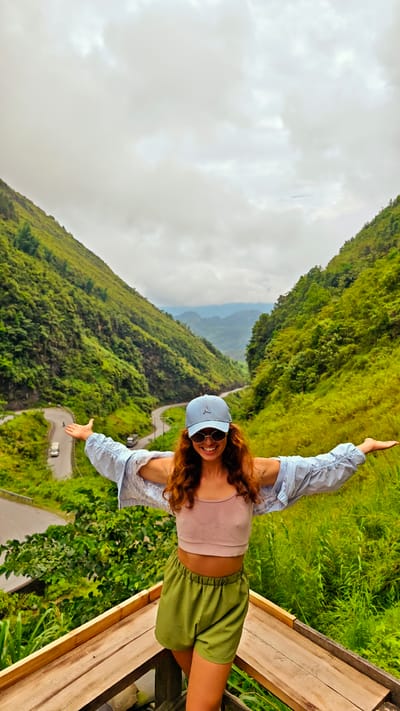 A woman stands on a wooden platform with her arms outstretched, smiling at the camera. She wears a blue baseball cap, sunglasses, a pink crop top, a light blue unbuttoned shirt, and green shorts. The background features a valley with lush green mountains and a winding road. The sky is cloudy. This is likely in the Phố Bảng Commune, Vietnam, given the landscape.