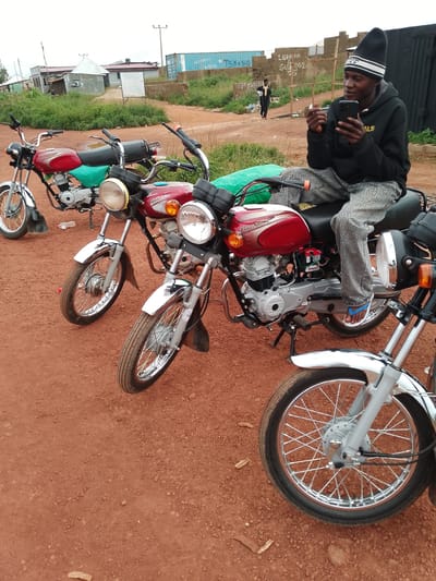 The media file depicts an outdoor scene in Agwala, Nigeria, with a focus on motorcycles and a young man interacting with his smartphone. Three motorcycles are visible, parked on a reddish-brown dirt ground. The motorcycles appear to be of a similar model, with red fuel tanks and silver fenders. One motorcycle is in the foreground, its front wheel and part of the engine prominently displayed. Another motorcycle is parked behind it, partially obscured. A third motorcycle, further in the background, is mostly red with green material draped over its seat.

A young man, wearing a black beanie with white stripes and a black hooded sweatshirt, is seated on one of the motorcycles. He is holding a smartphone in his hands and appears to be looking at it. He is wearing grey, patterned trousers and blue and white flip-flops. His posture suggests casual engagement with his device.

In the background, there are buildings with corrugated metal roofs and some brick structures. A large, light blue shipping container is visible to the left, and other containers or structures are present behind a brick wall. There is also vegetation growing in the area. A second person, wearing a dark robe and a yellow garment, is walking away from the camera in the background, adding a sense of everyday activity.

The sky appears overcast, suggesting a cloudy or possibly rainy day. The overall lighting is diffused. The scene captures a moment of everyday life, with transportation (motorcycles) and personal technology (smartphone) playing a central role. The presence of multiple motorcycles hints at a transportation hub or a gathering of individuals who use these vehicles. The setting appears to be a semi-urban or rural area, given the mix of buildings, open space, and dirt ground. There is some visible text on the brick wall in the background, but it is largely illegible.