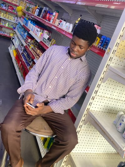 An individual is depicted seated on a brown, cushioned stool within a retail store environment, possibly located in Kabala, Nigeria. The subject is a male with dark skin and short, dark hair, wearing a purple and white checkered long-sleeved shirt and brown trousers. He is holding a white smartphone in both hands and looking down at the device. His bare feet are visible. The background consists of multiple white metal shelving units with red trim, stocked with various consumer products, primarily toiletries, personal care items, and other packaged goods. Specific items include bottles of shampoo, lotions, and deodorants. Some shelves appear sparsely populated. A sign partially reading "PLEASE DON'T SPRAY" is visible on a shelf above the individual's head. The store appears to be lit by artificial lighting.