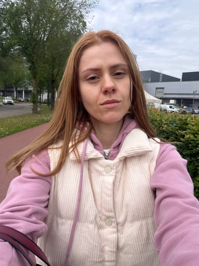 A woman with long, auburn hair takes a selfie outdoors. She is wearing a pink hoodie layered under a cream-colored corduroy vest with large white buttons and a purple drawstring. She is holding a dark red purse strap with her left hand. The background shows a paved path, trees, a street with cars, a building with a large white tent in front of it, and a dense green hedge. The scene is consistent with a suburban or commercial area in Son en Breugel, Netherlands, likely near a road or pathway suitable for pedestrians. The lighting suggests an overcast day.