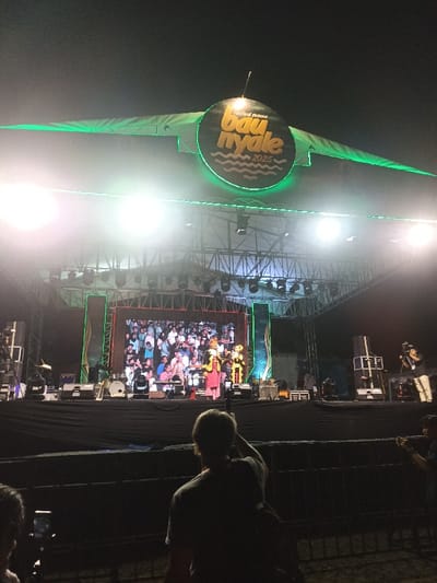 This nighttime image captures a vibrant outdoor stage setup during what appears to be a festival or concert in Kecamatan Pujut, Indonesia. A large, circular sign prominently hangs above the stage, featuring green neon lighting and the text "festival pesona bau nyale 2025" in yellow lettering, accompanied by stylized wave graphics.
The stage itself is well-lit by numerous bright spotlights, creating visible lens flares. On the stage, a performance is in progress, featuring two distinct figures that resemble puppets or mascots – one in red and yellow attire, and another in yellow and black. A person is visible between these figures, potentially a performer or presenter. Behind them, a large LED screen displays an image of a crowd, possibly a live feed of the audience or a pre-recorded segment. Musical instruments, including a drum set, are arranged on the stage. To the right, a cameraman can be seen, capturing the event.
In the foreground, several audience members are visible, mostly silhouetted against the bright stage lights. One person in the immediate foreground, with their back to the camera, is holding up a smartphone, actively recording the performance. Other attendees also appear to be holding up their phones, indicating engagement with the event. The dark sky confirms the activity is taking place at night. The visible text on the sign, "Bau Nyale 2025," suggests this is the annual Bau Nyale Festival, a significant cultural event in Lombok, Indonesia.