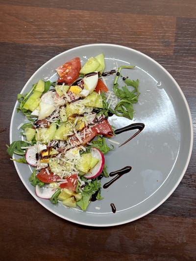 A close-up, overhead shot showcases a grey plate of salad placed on a dark wooden table. The salad is composed of arugula, sliced radishes, cherry tomatoes, cucumber, hard-boiled egg, and shredded parmesan cheese. A balsamic glaze is drizzled over the salad and onto the plate. The scene is set in Balbriggan, Ireland.