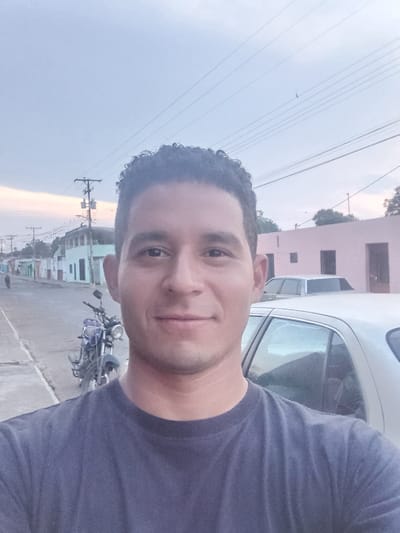 A man is taking a selfie. He is wearing a dark blue t-shirt. He is standing next to a white car. In the background, there is a street with a parked motorcycle, buildings painted in various colors, and power lines. The image appears to be taken in the city of Duaca, Venezuela. The sky is a light color, possibly indicating dawn or dusk.