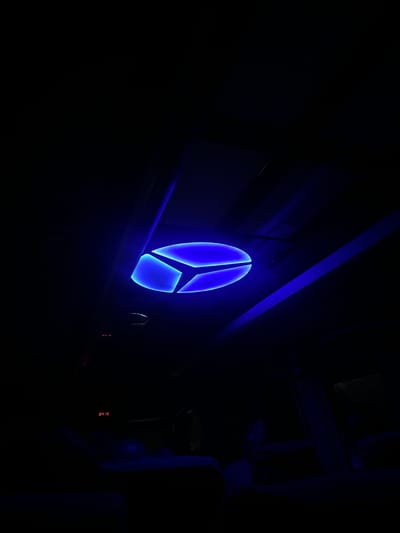 The scene depicts the interior of a darkened space, located in the city of Khashuri, Georgia. A prominent blue, oval-shaped light fixture, divided into three illuminated segments, is mounted on the ceiling in the upper center of the frame. This fixture is the primary light source, casting a deep blue hue across the visible ceiling panels and structural elements. The majority of the lower and peripheral areas of the image remain in profound darkness due to the low light conditions. In the lower-left quadrant, a faint red digital display indicates "04:16," with additional small red indicator lights visible above it. Indistinct dark forms in the lower foreground suggest the presence of occupied seats or individuals within the dimly lit environment.