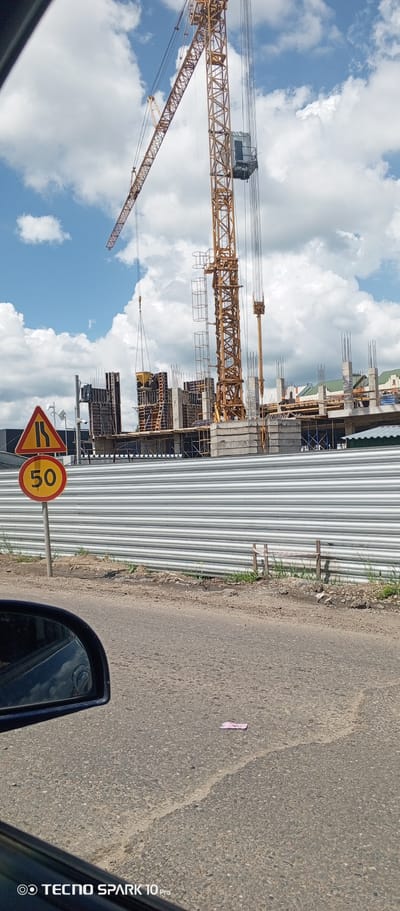The image depicts a construction site with a large yellow crane in the background. A partially constructed building is visible behind a corrugated metal fence. In the foreground, a road sign indicates a narrowing road ahead and a speed limit of 50. The scene is likely in an urban area, possibly Bereg Kubani, Russia. The sky is partly cloudy. A portion of a car's side mirror is visible in the lower left corner, suggesting the image was taken from inside a vehicle. A small piece of paper is on the road.