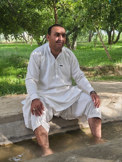 The image features a man sitting with his feet submerged in a narrow channel of water, possibly for cooling. He is wearing a white shalwar kameez, a traditional South Asian garment. His right hand, adorned with a ring, rests on his left leg. A bracelet is visible on his left wrist. The man is positioned outdoors, with lush green grass and trees forming the background. The sunlight suggests it is daytime. The setting appears to be a rural or natural environment, consistent with the provided location context of Kan Mehtarzai, Pakistan. The man's expression is neutral, and he is looking towards the camera. There is a visible tag on his shirt that appears to have text on it.