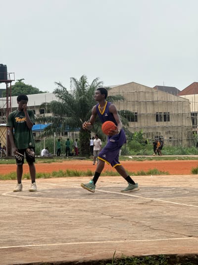 The image shows an outdoor basketball court with a dirt and concrete surface. Two young Black men are the primary subjects. One, wearing a purple and yellow basketball jersey and shorts, is dribbling an orange basketball. He is in mid-motion, with his body leaning forward. The other man, wearing a green t-shirt and black shorts with white lettering, stands on the court, appearing to observe the game. Several other people are visible in the background, some appearing to be playing basketball or watching. In the background, there is a partially constructed building with scaffolding, palm trees, and other greenery. The sky is overcast, suggesting a cloudy or potentially rainy day. The overall scene depicts a casual basketball game or practice taking place in an urban or semi-urban environment in Amansee, Nigeria. There is some visible text on the green t-shirt worn by one of the subjects, and on the shorts worn by the same subject. The text on the shorts is partially obscured but appears to say "NVAN".