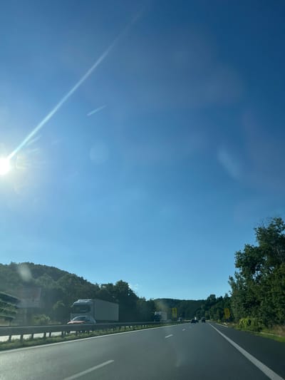 The image captures a view from a vehicle traveling on a highway in the vicinity of Dolna Vasilitsa, Bulgaria. The sky is a clear, bright blue, with the sun prominently visible in the upper left corner, casting a strong glare and lens flares across the upper portion of the image. The highway has multiple lanes, marked with white lines, and a metal guardrail runs along the side.
On the highway, there are several vehicles, including a large white truck with a trailer in the left lane and a smaller orange car in front of it. Further ahead, other vehicles, including cars and possibly trucks, are visible in the distance on the highway. The road is flanked by lush green trees and rolling hills covered in dense foliage. There are also some road signs visible, including yellow warning signs on the right side of the road. A large billboard is partially visible on the left side, further back, behind the guardrail and trees. The overall scene depicts a typical highway landscape on a sunny day, likely in a rural or semi-rural area given the surrounding vegetation and topography.