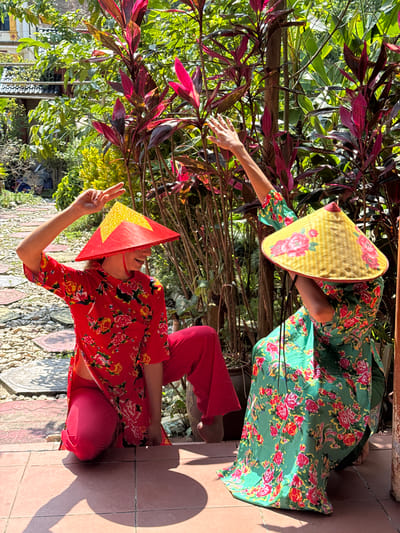 This vibrant outdoor photograph captures two individuals, likely women, posing amidst lush greenery and brightly colored plants. The scene is illuminated by strong sunlight, creating distinct shadows on the terracotta-tiled ground, suggesting a clear, sunny day.

The person on the left is standing and wears a traditional Vietnamese conical hat, or nón lá, which is yellow with delicate pink floral patterns. They are dressed in a flowing green garment adorned with prominent red floral designs. Their right arm is extended forward and slightly upward, as if reaching out or gesturing.

The second individual, positioned on the right, is in a squatting or kneeling pose. They wear a red outfit with a matching floral print top and red pants. Their red conical hat is striking, featuring a large yellow five-pointed star, reminiscent of the Vietnamese flag. This person is holding up their left hand, making a peace sign. Both individuals' faces are mostly obscured by their hats or turned away from the camera.

The background is rich with foliage, including tall plants with dark green leaves and bushes with striking maroon-red leaves, contributing to a vibrant tropical or subtropical aesthetic. The ground consists of reddish-brown tiles and a patterned section made of small, light-colored stones. The traditional attire, particularly the nón lá with the national star, strongly places the scene within Vietnam, consistent with the Thông Nguyên Commune location context. No visible text is present.