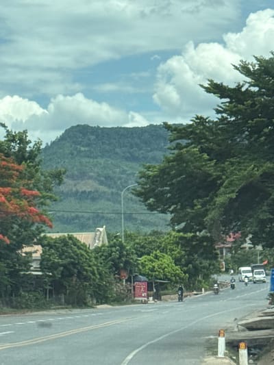 This is a landscape image taken from a roadside perspective, oriented vertically. The scene is dominated by a lush green, tree-covered mountainside in the background. In the foreground, a paved road with yellow dividing lines stretches into the distance. To the left of the road, there are trees and a few buildings, including what appears to be a light-colored structure with a pitched roof. A lamppost is also visible.
Several motorcycles and a white van are present on the road, suggesting vehicular traffic. Some people can be seen on or near the motorcycles, and one person stands on the side of the road. The sky is partially obscured by large, white clouds against a light blue background. The presence of greenery and the types of vehicles suggest a rural or semi-rural setting, likely in a tropical or subtropical climate given the lush vegetation. The time of day appears to be daytime, with good natural lighting.
There is some visible text on what looks like a red sign on the left side of the road, but it is blurry and unreadable. The overall impression is a snapshot of daily life on a road in a mountainous region, possibly in Southeast Asia.