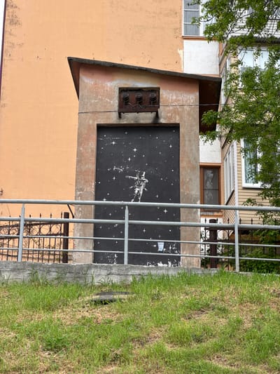 The image shows a section of a building with an adjacent grassy area. A metal fence partially obscures the view. On the building, there's an abstract mural painted on a recessed black rectangular area. The mural appears to depict an astronaut or figure in space amongst stars. Above the mural, there's a rusty electrical box with exposed wires. The building is painted in shades of orange and tan. The upper floors of the building, visible above the mural, show windows and different architectural styles. To the right of the building is a patch of green grass. The image provides no information about Zamankul, Russia, or any people, actions, or interactions at the location.