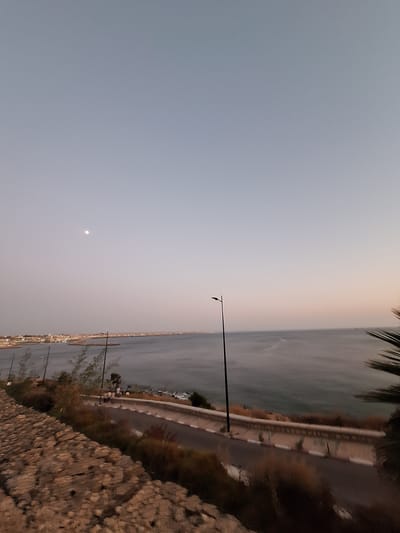 This is a vertical, low-angle shot of a coastal scene at dusk. The sky takes up the left two-thirds of the frame, fading from a pale, hazy blue at the top to a soft, peachy orange near the horizon. A small, bright circle, likely the moon, is visible in the upper left quadrant of the sky. The right third of the image depicts the sea and a road running along the coast. The sea is a deep, dark blue, reflecting the dimming light. A road with white markings and streetlights runs alongside a low wall and then a rocky embankment. Palm fronds are visible in the upper right corner, suggesting a tropical or subtropical location. The overall mood is peaceful and serene, with the fading light and calm sea creating a sense of tranquility. The scene appears to be a seaside promenade or a road overlooking the ocean, possibly in Safi, Morocco, given the location context. The time of day is clearly dusk, as indicated by the soft sky colors and the presence of streetlights. No people are distinctly visible, but there are faint signs of activity along the road and in the distance. The rocky embankment in the foreground is textured and rough, adding a natural element to the composed scene.