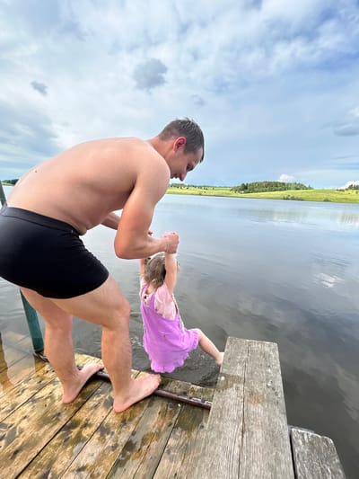 In this photograph, a shirtless man wearing black swimming trunks is on a wooden dock. He is holding the hands of a young girl who is wearing a purple dress. The man is facing away from the camera, looking towards the water and the girl. The girl is upside down, with her legs in the air, as if she is being swung. The wooden dock extends into a body of water, which reflects the sky and a green, tree-lined landscape in the distance. The sky is cloudy, suggesting it might be overcast or a partly cloudy day. The scene appears to be a leisurely summer day at a lake or river in a rural setting, possibly in Верх-Рождество, Russia. The man's posture and the girl's playful inversion suggest a lighthearted and fun moment between them. There is no visible text in the image.