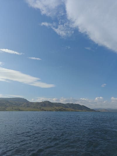 The image depicts a scenic view of a body of water, likely a lake, with rolling hills and mountains in the background. The sky is a clear blue with scattered white clouds. The water is dark blue and appears to be slightly choppy. The hills in the distance are covered with green vegetation and some scattered trees. The overall scene is peaceful and natural. The image does not contain any people or buildings, and therefore it is impossible to determine the specific location in the city of Manat, Kazakhstan.
